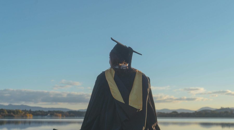 Back View of a Student Wearing a Graduation Gown and a Mortarboard, Sitting by a Lake